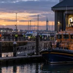 Illustration of a lifeboat station at Portsmouth harbour with a rescue vessel alongside while police monitor a crowd across the marina.