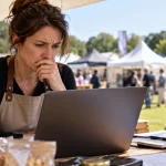 Editorial illustration of a food-festival trader looking concerned while reading a laptop at an outdoor market stall.